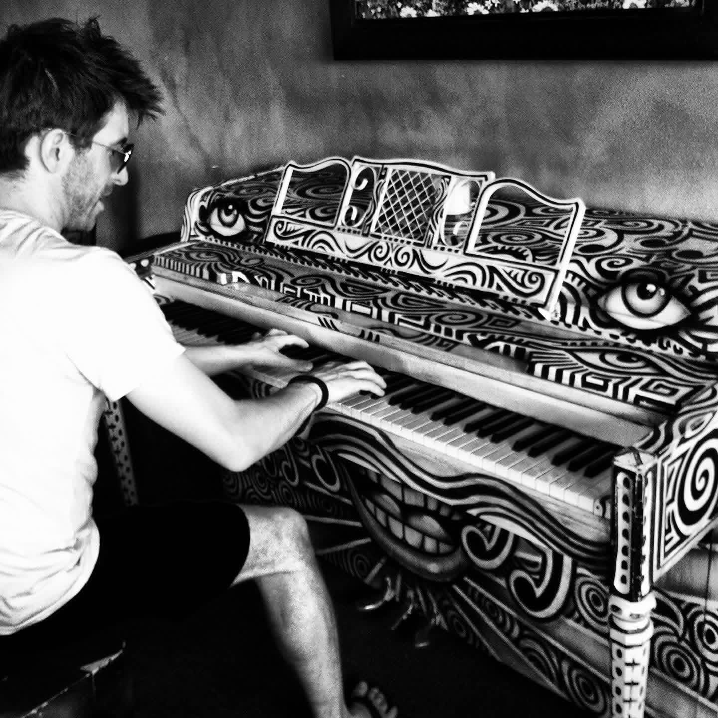 Chris LoPresto in sunglasses playing a hand-painted upright piano covered in op-art eyes and swirls
