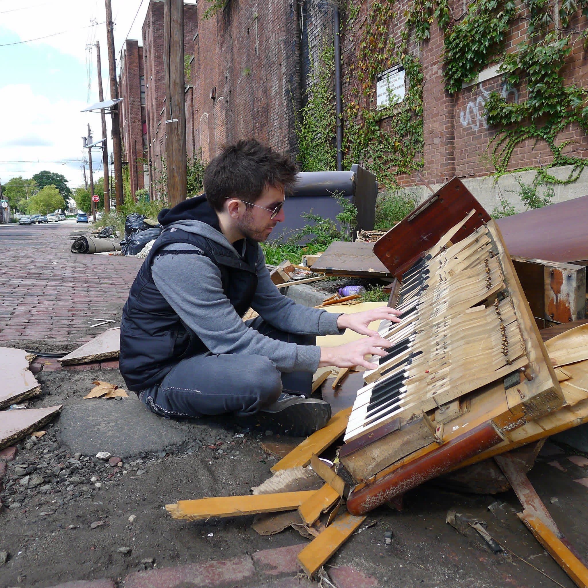 Chris LoPresto sitting cross-legged on a brick street playing a wrecked upright piano